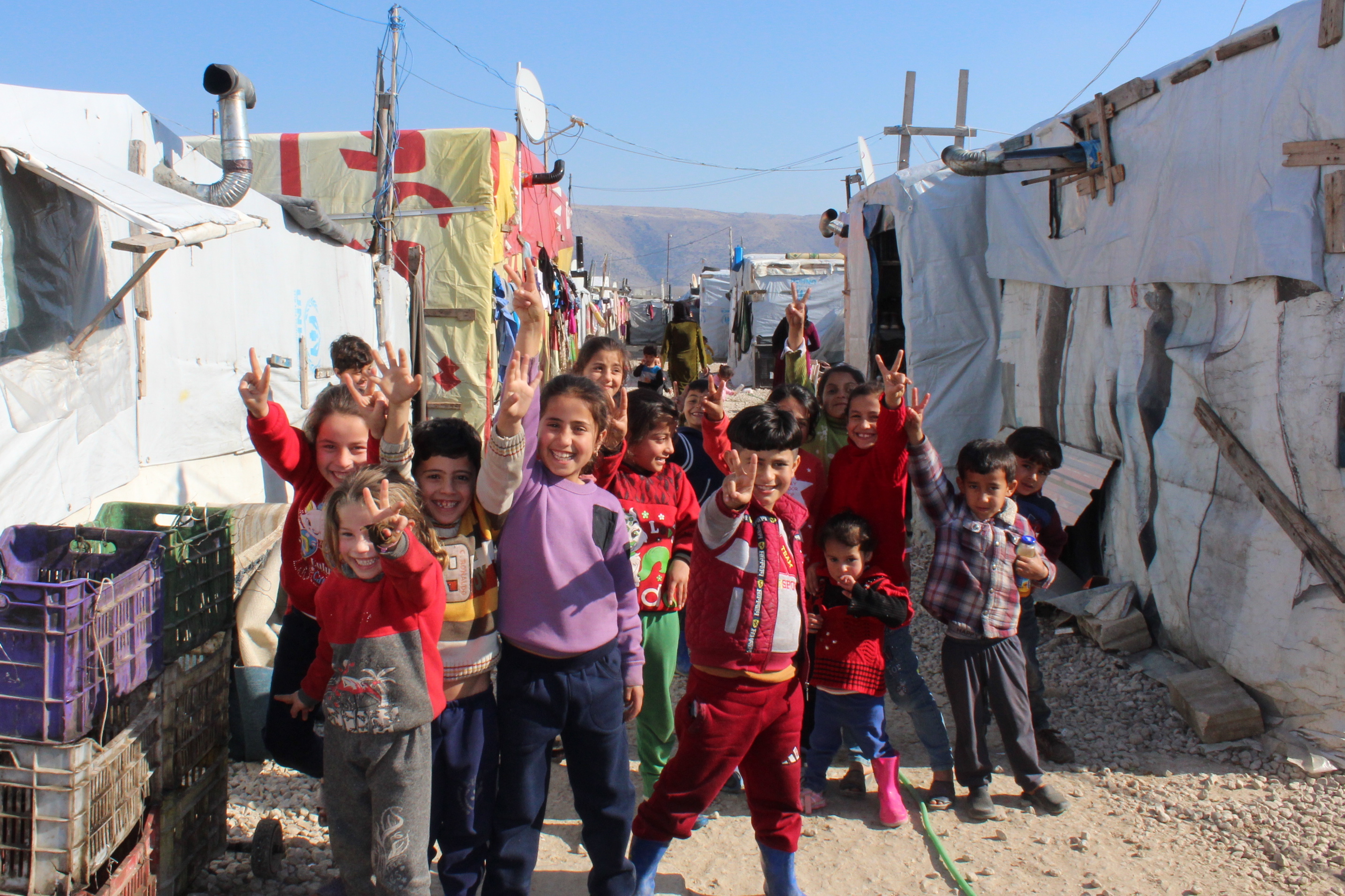 eine Gruppe Kinder im Grundschulalter grinst in die Kamera, im Hintergrund sind Zelte eines Flüchtlingscamps erkennbar