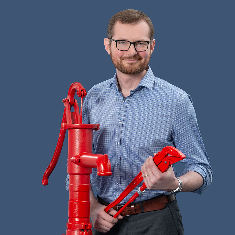 Mathias Anderson steht lächelnd neben einer roten Pumpe und hält eine große rote Rohrzange in der Hand